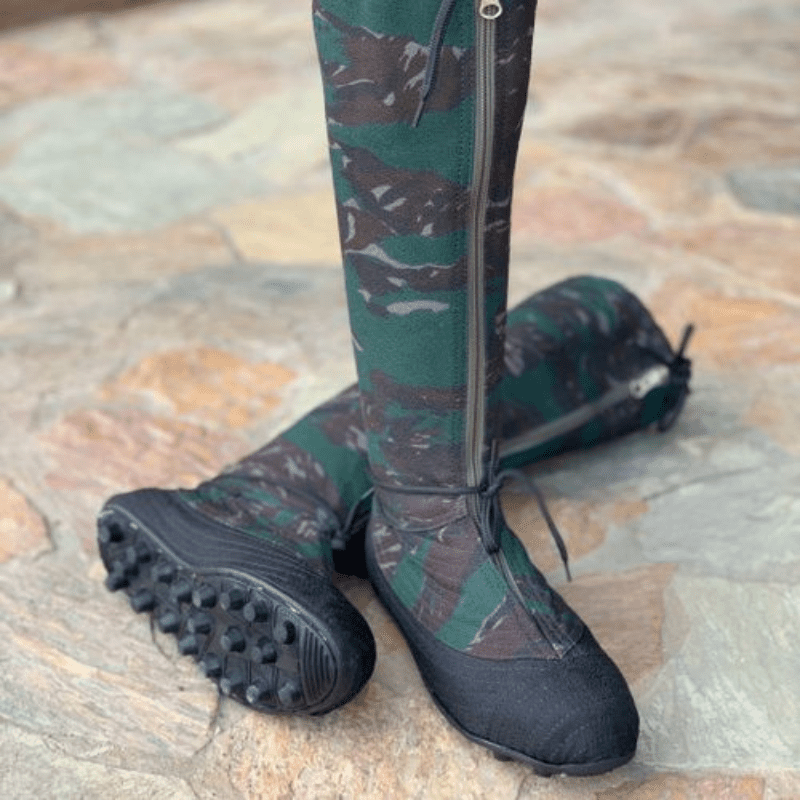The image shows a pair of tall, camouflage-patterned boots. The boots are predominantly dark green and brown, with a camouflage pattern. They have black soles with small studs and appear to be made of a fabric material. A zipper is visible on the side of each boot. The boots are positioned on a stone or paved surface.